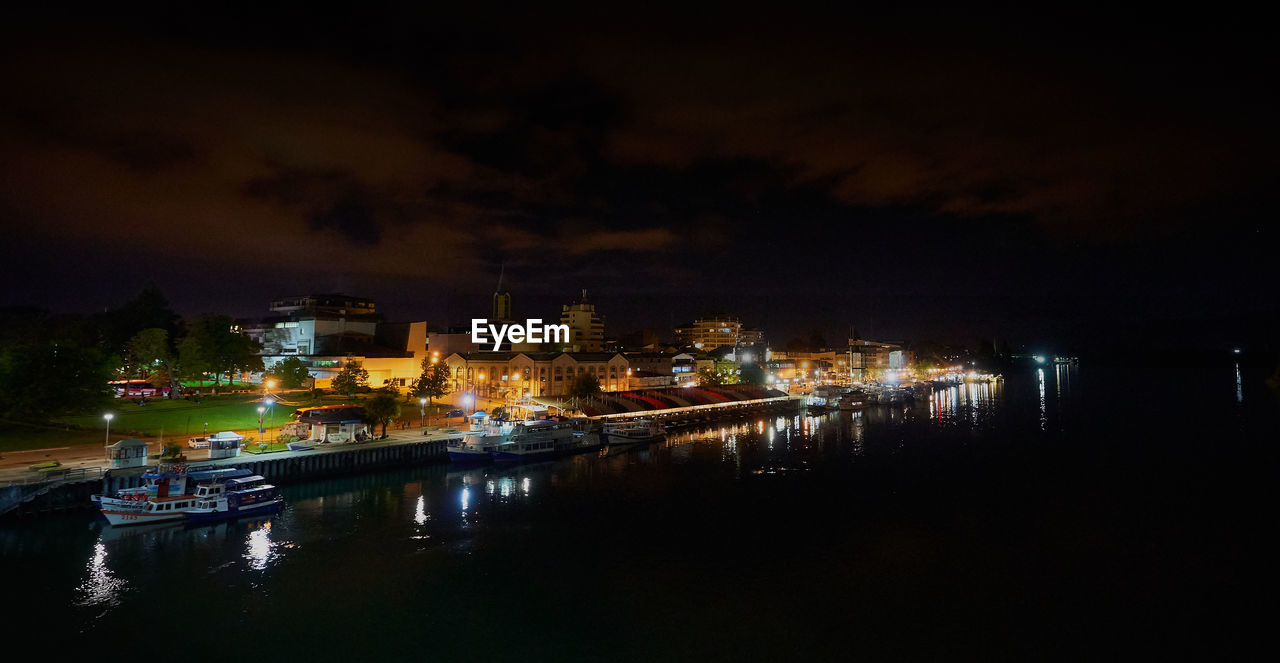 Illuminated buildings by river against sky at night