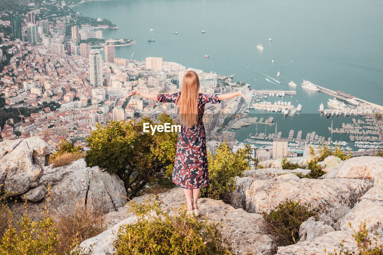 Young woman with long hair in dress on cliff. panoramic view of monaco, sea coast, french riviera.