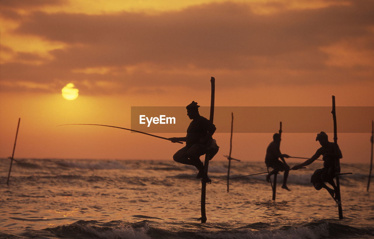 Side view of silhouette man and woman fishing in sea from wooden posts at sunset