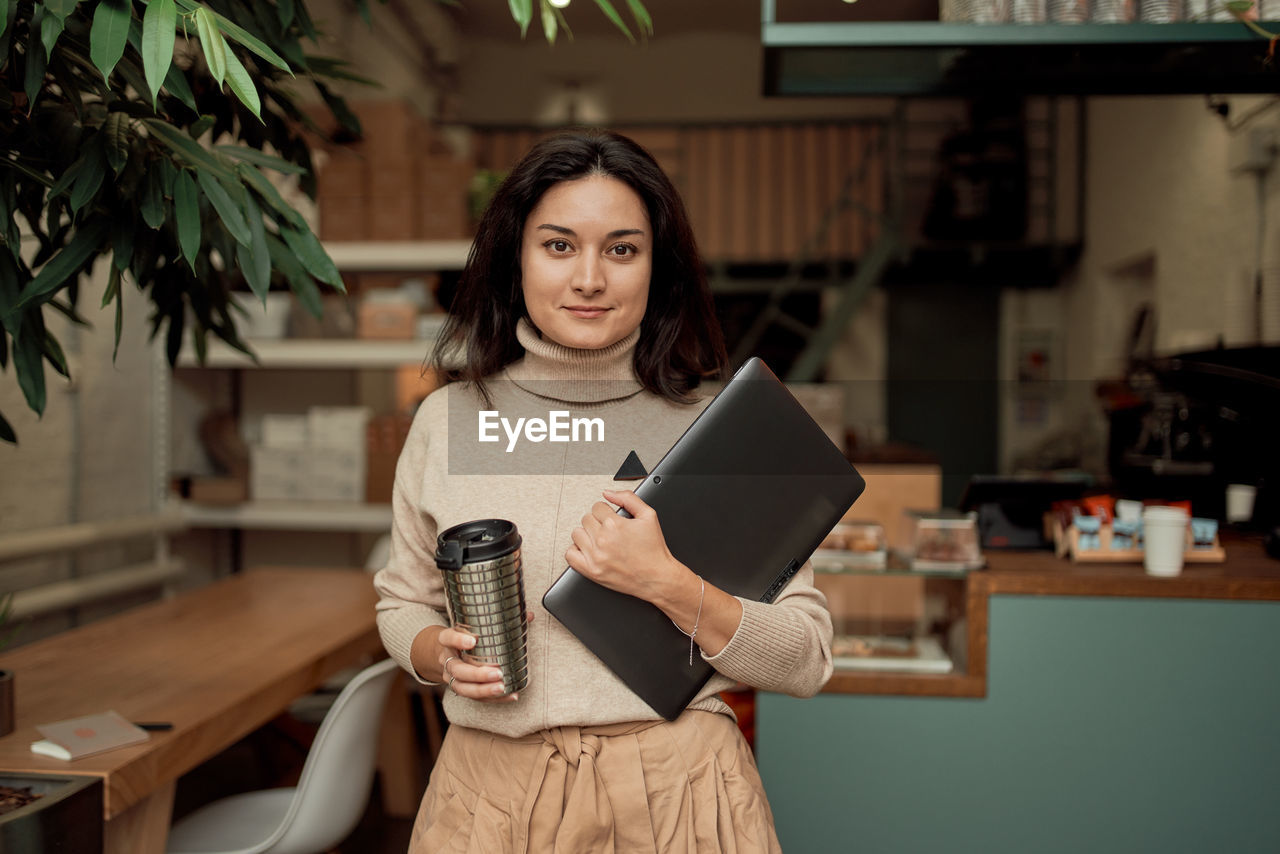 Portrait of businesswoman holding coffee cup and laptop at cafe