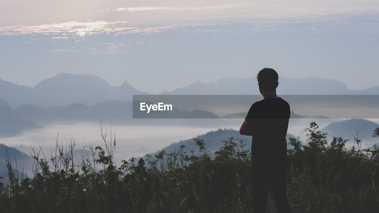 Rear view of silhouette man standing on mountain against cloudy sky