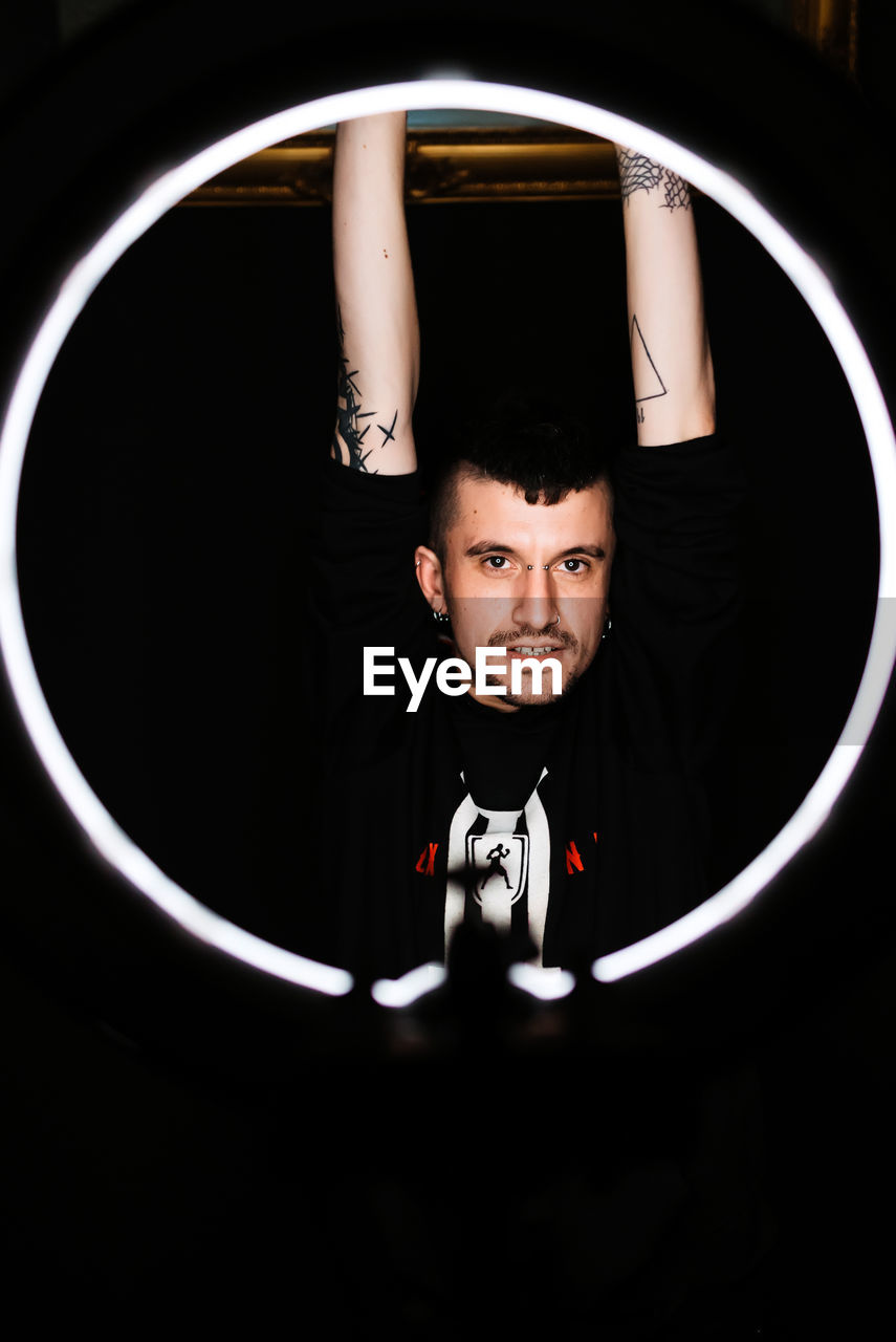 Trendy tattooed guy with raised arms looking at camera while standing behind round illumination in dark room