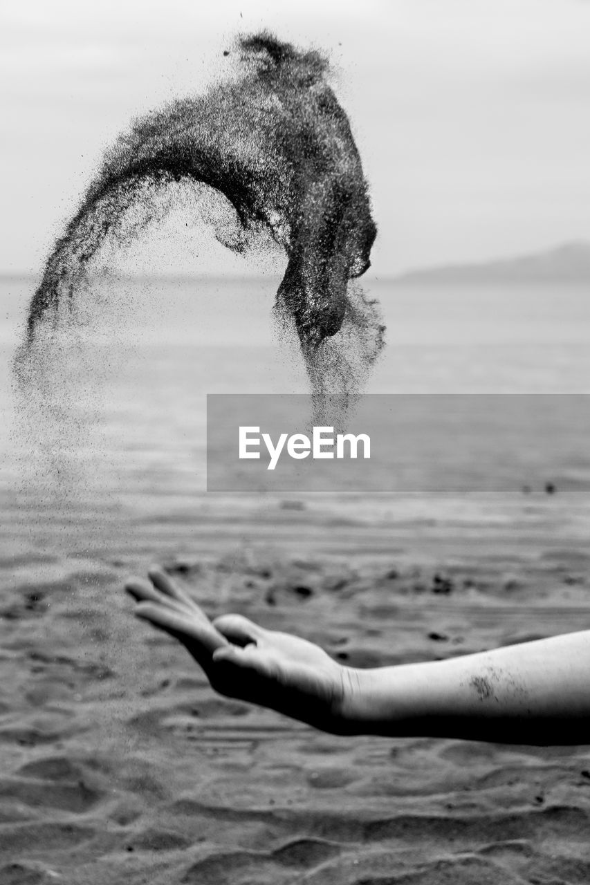 Cropped image of hand throwing sand at beach against sky