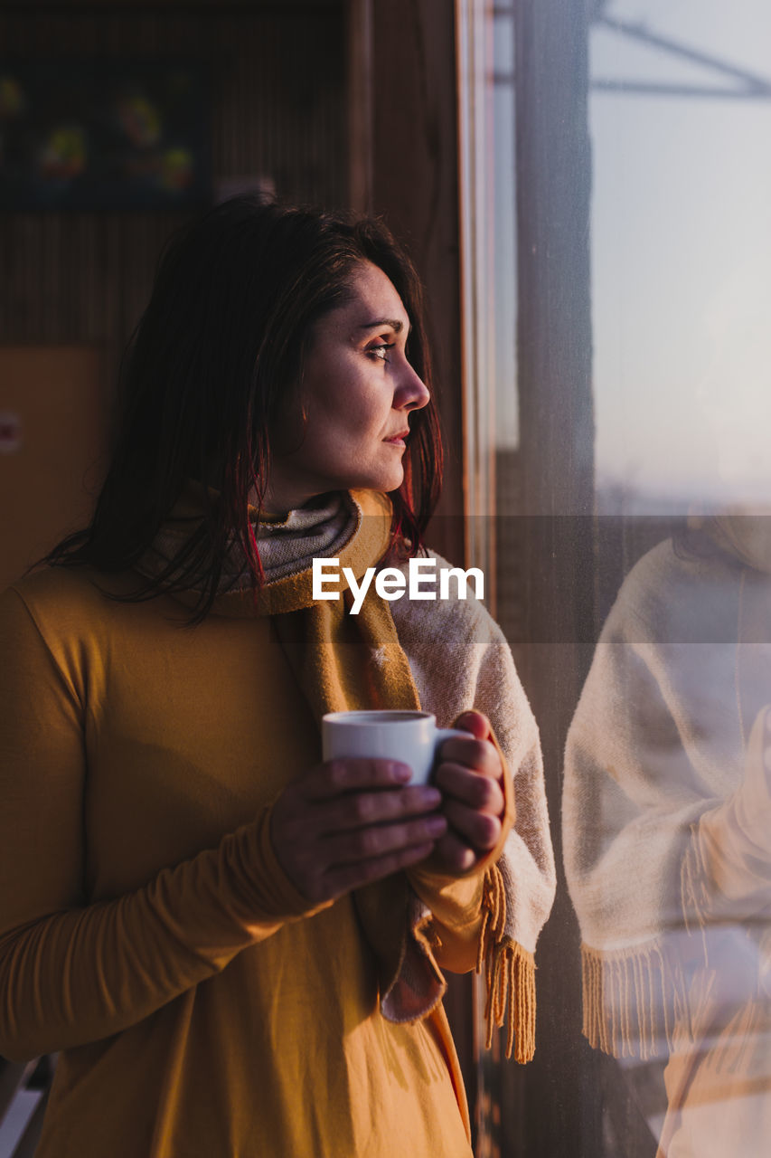 Woman with coffee looking towards window while standing at home