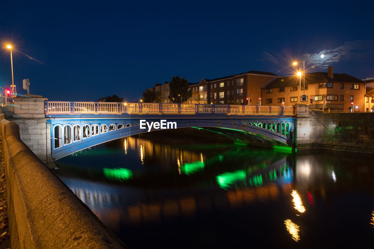 Illuminated bridge over river by buildings against sky at night