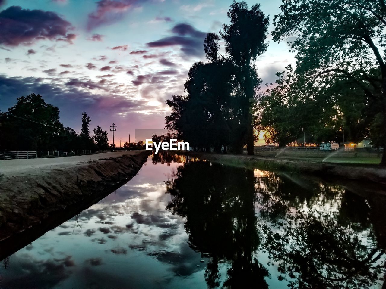 REFLECTION OF SILHOUETTE TREES IN LAKE AT SUNSET