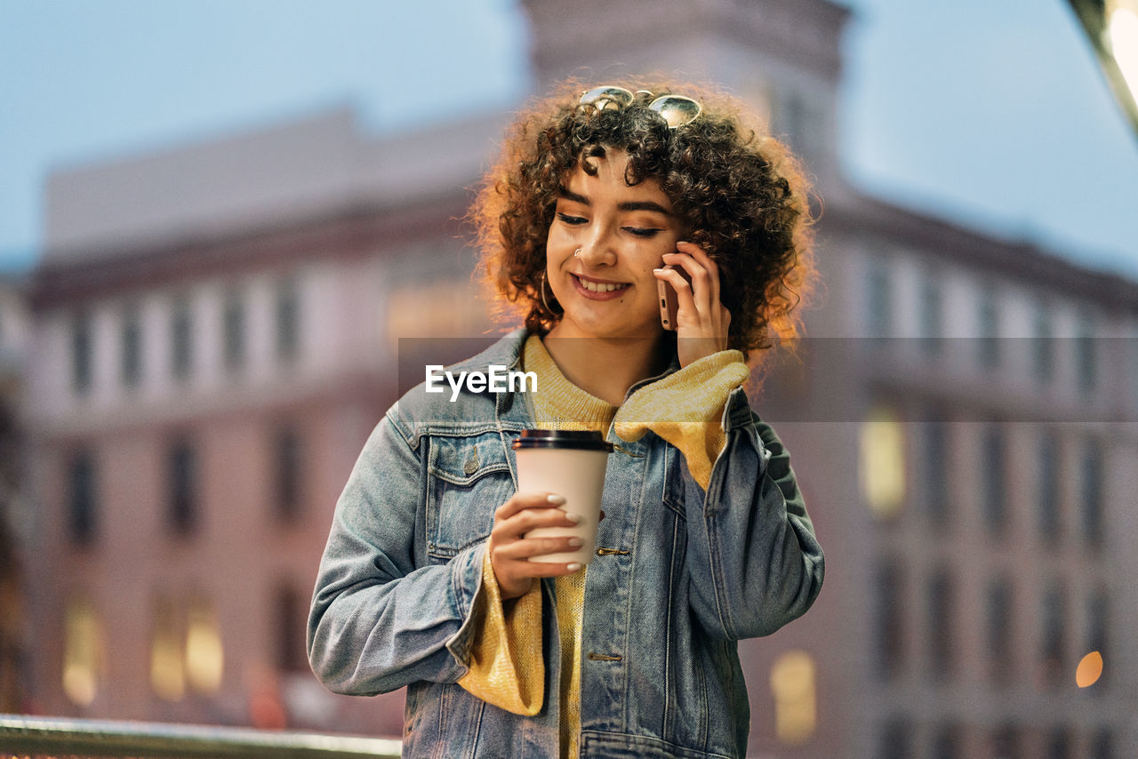 Young woman holding coffee cup talking on phone against building