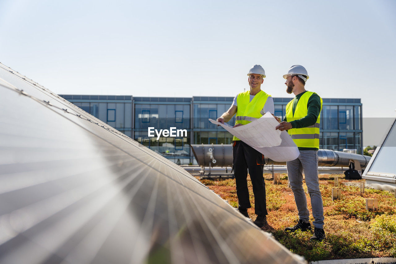 Two technicians analyzing blueprints on the rooftop of a corporate building equipped with solar panels