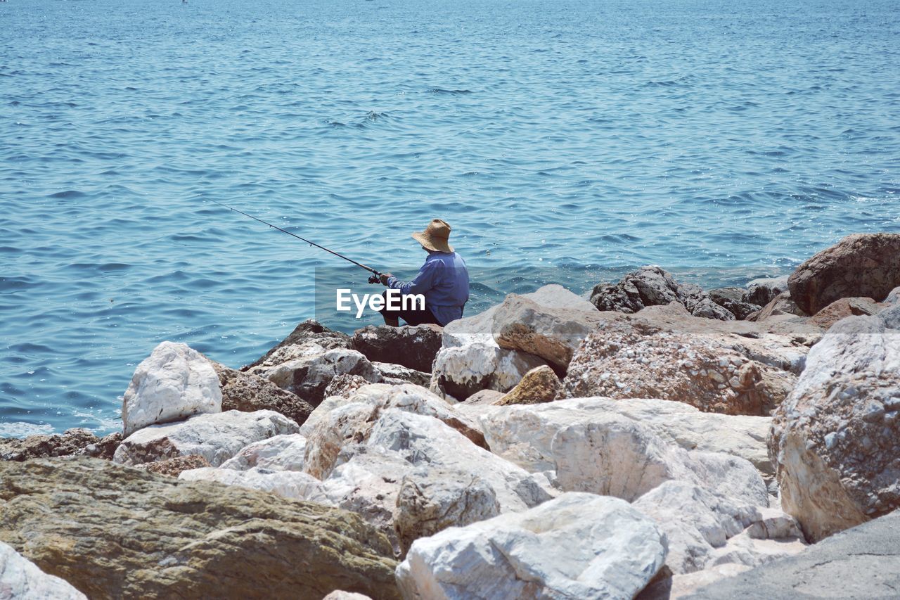 Side view of man fishing while sitting on rock at sea