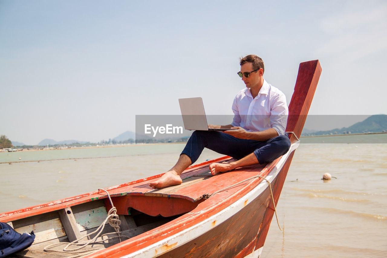 Side view of business man working with laptop on boat on sea against clear sky