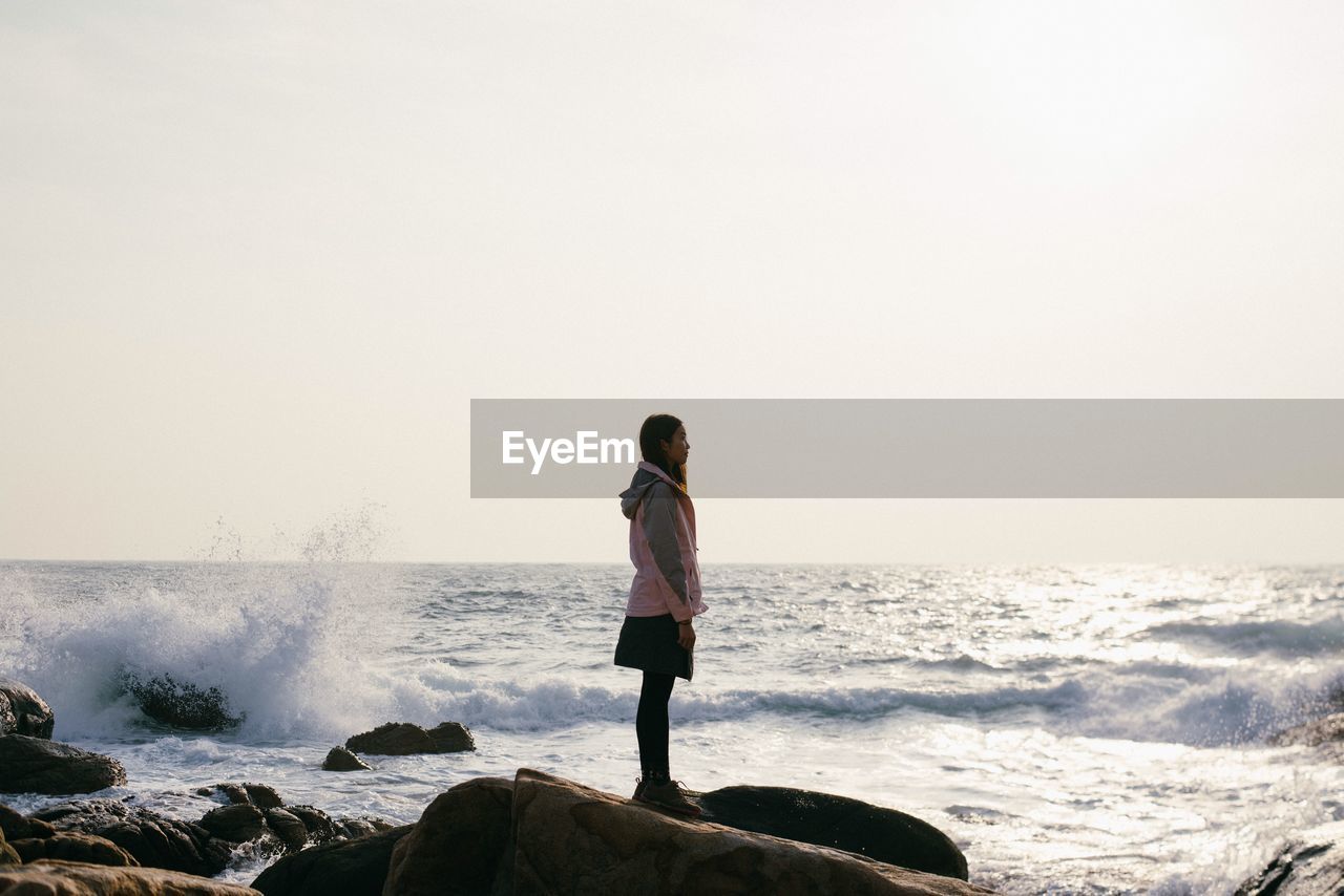 Side view of young woman standing on rock against splashing sea waves