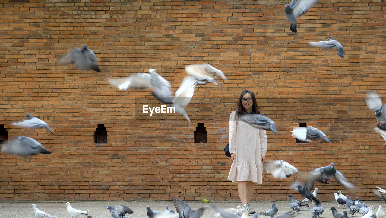 Woman standing by flying birds against brick wall