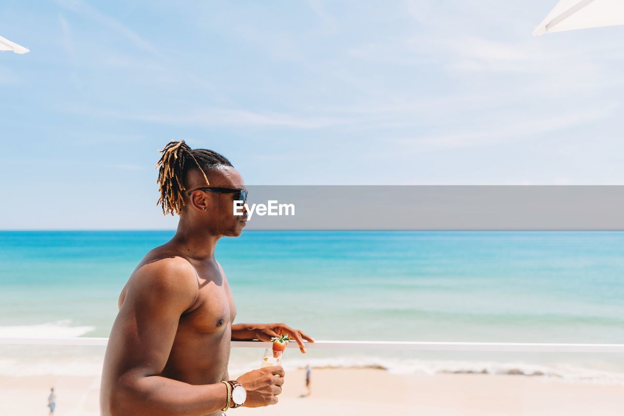 Shirtless young man wearing sunglasses while holding drink at beach against sky