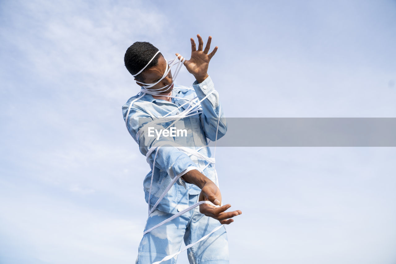 Young man wearing cloud patterned denim outfit struggling to get free from rope while standing against sky