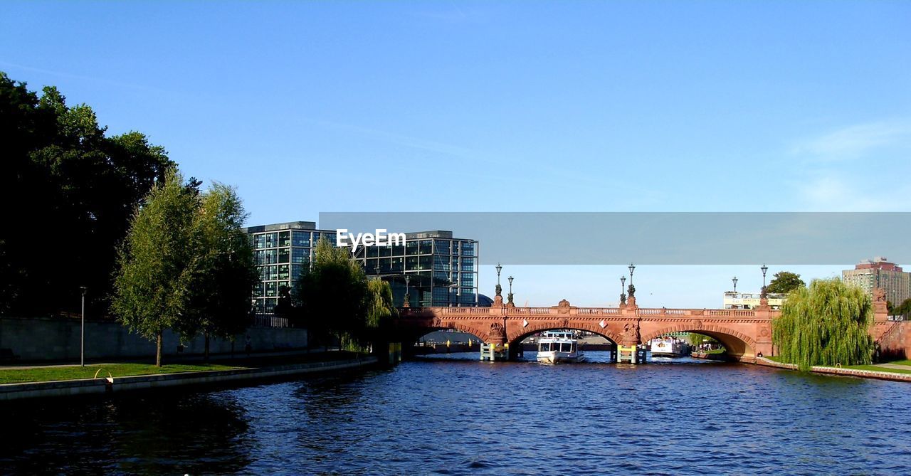 architecture, built structure, water, river, bridge, reflection, sky, tree, building exterior, waterway, nature, plant, city, body of water, travel destinations, waterfront, transportation, blue, no people, cityscape, building, travel, day, clear sky, outdoors, tourism, arch bridge