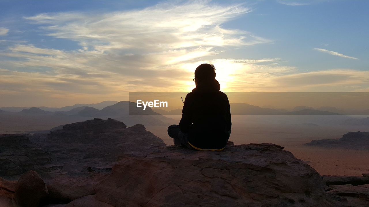 Boy sitting on rock formation against sky during sunset
