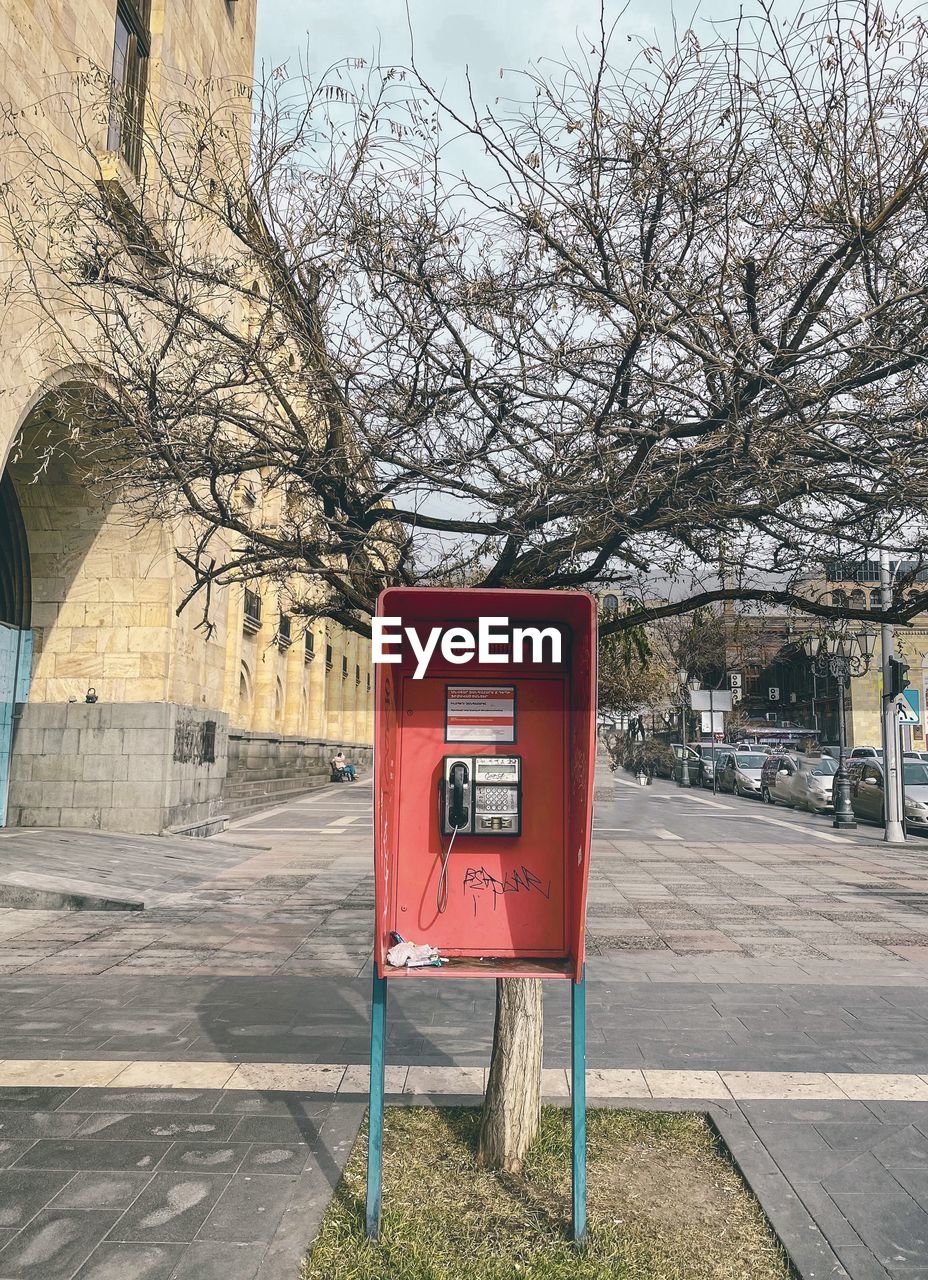 city, tree, communication, urban area, plant, architecture, telephone, nature, bare tree, telephone booth, day, built structure, mail, no people, mailbox, building exterior, payphone, street, text, branch, post box, outdoors, road, public mailbox, correspondence, sign, red, convenience, technology, sky, lane, sidewalk