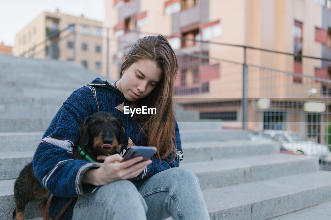 Young adult woman looking her smartphone sit next to her dachshund dog
