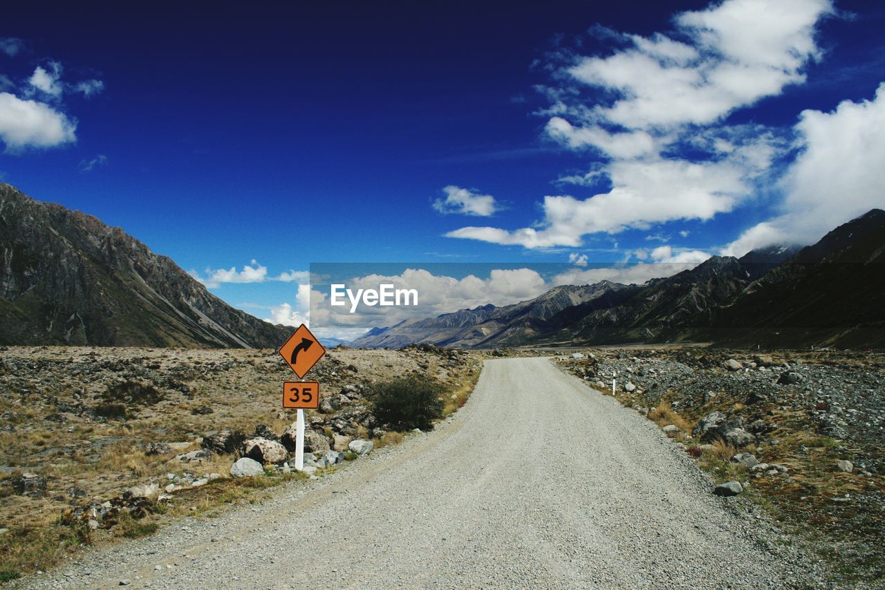 Empty road amidst field by mountains against cloudy sky