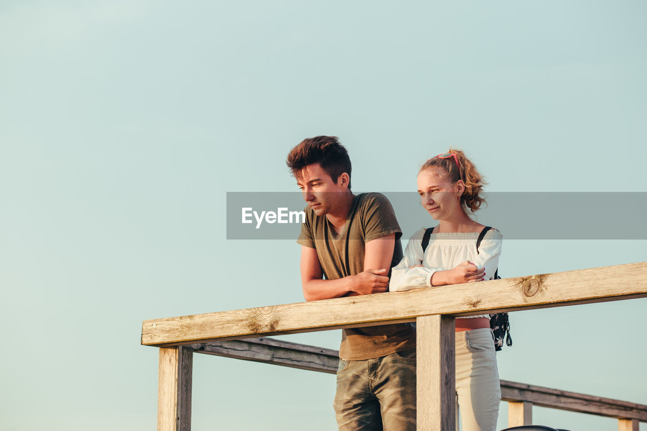 Low angle view of couple looking away while standing by railing against clear sky