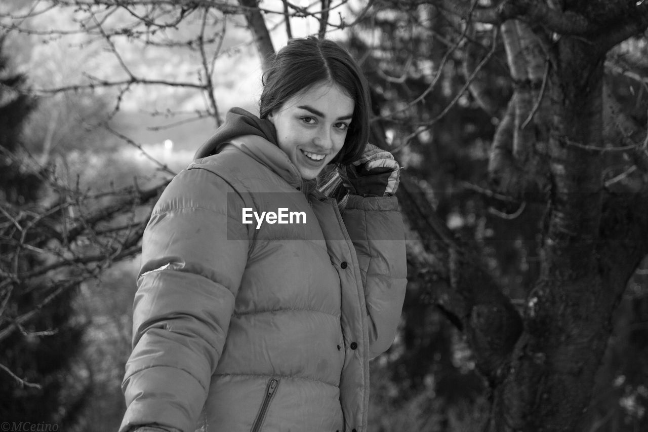 Portrait in black and white of smiling young woman standing against trees during winter 