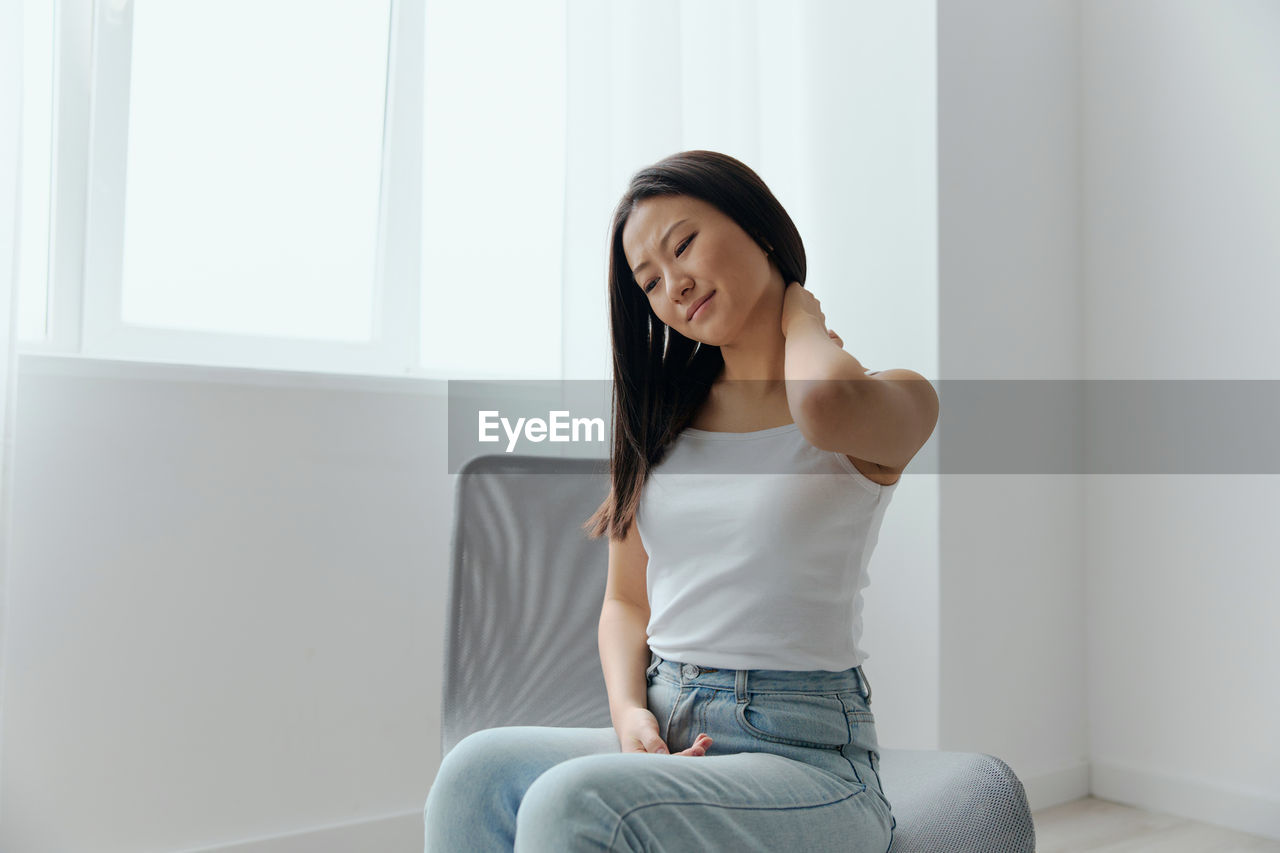 one person, adult, sitting, women, indoors, young adult, casual clothing, lifestyles, relaxation, clothing, emotion, three quarter length, long hair, front view, hairstyle, photo shoot, copy space, looking, person, domestic room, smiling, home interior, happiness, jeans, window, domestic life, limb, contemplation, female, brown hair, human leg, furniture, portrait, living room, day, technology, communication, full length