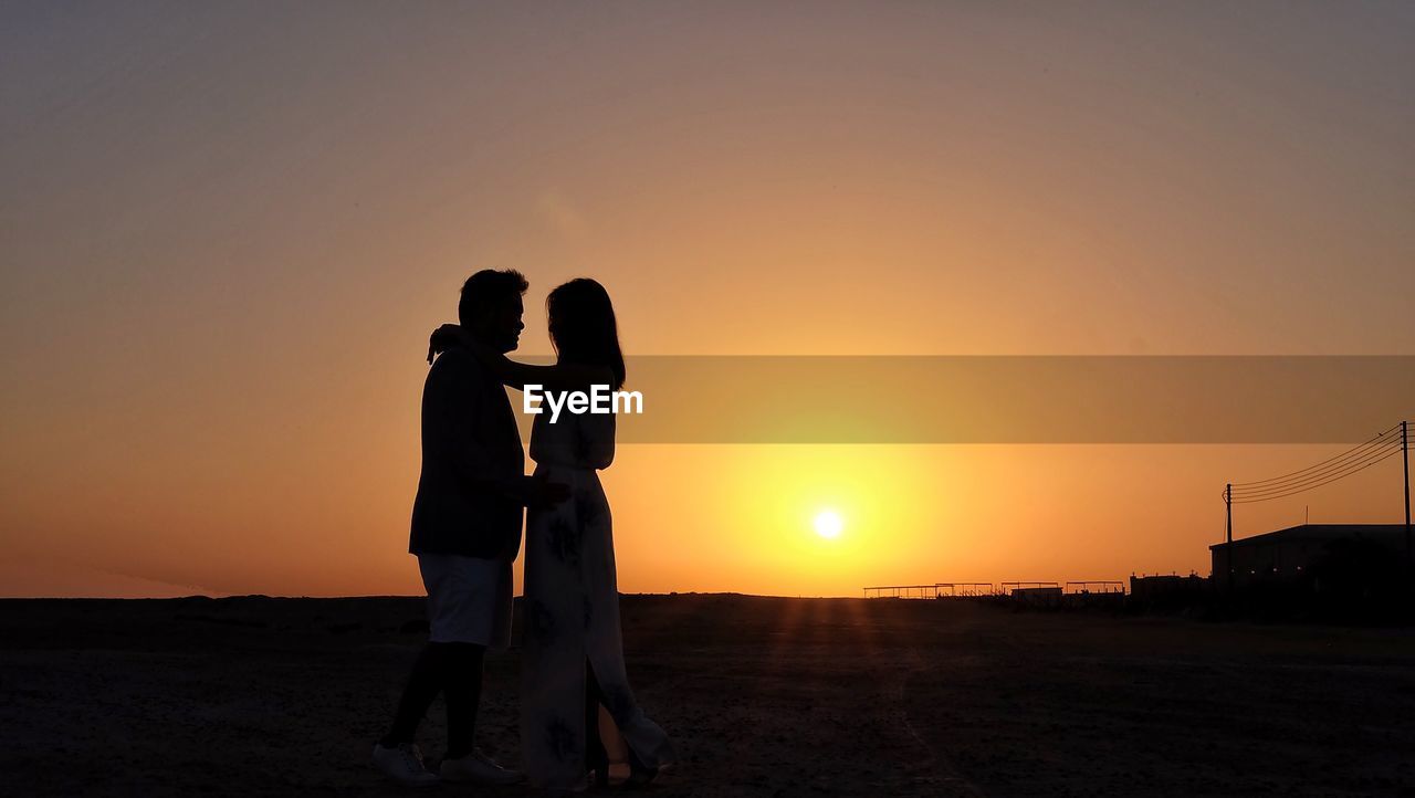 Side view of couple standing against orange sky during sunset