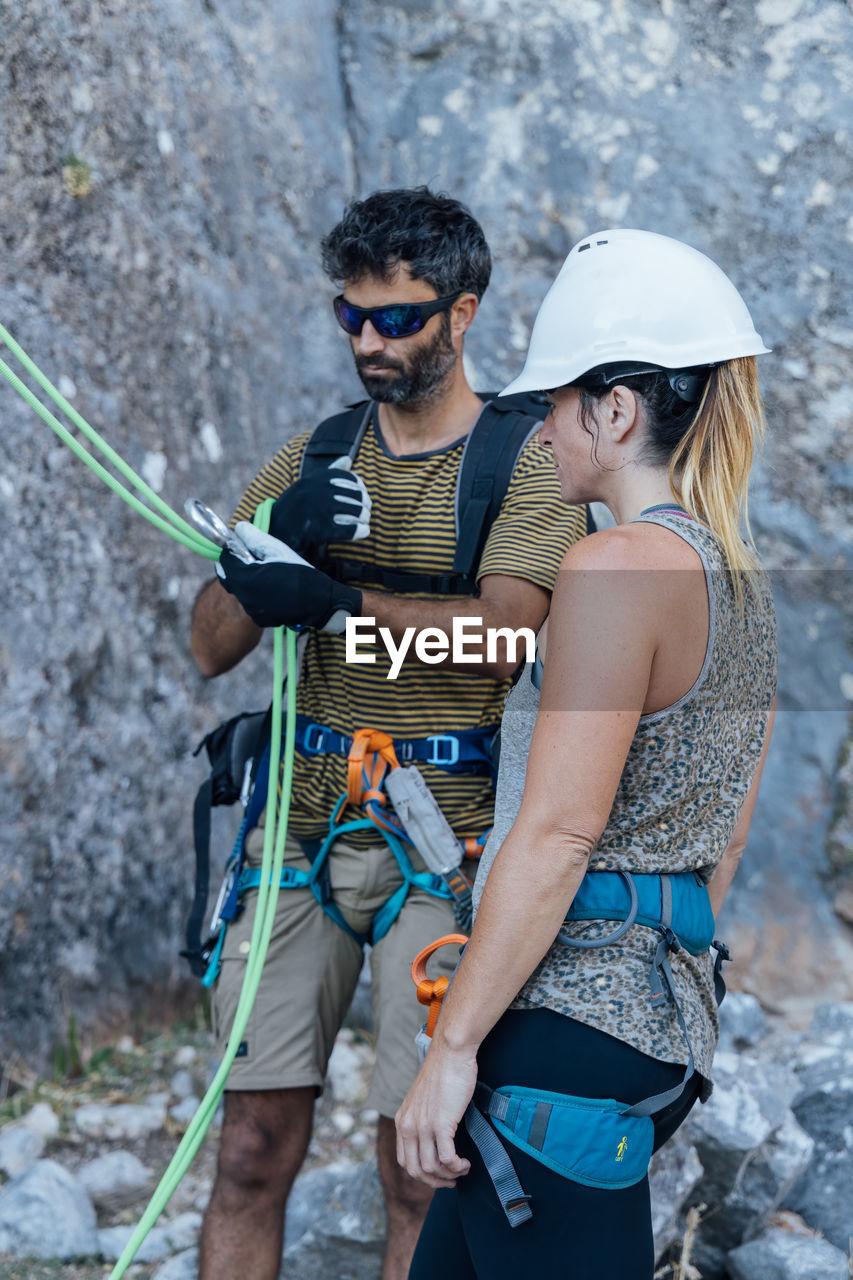 Fearless active couple in safety harnesses preparing gear for ascending rocky mountain slope while practicing climbing in nature