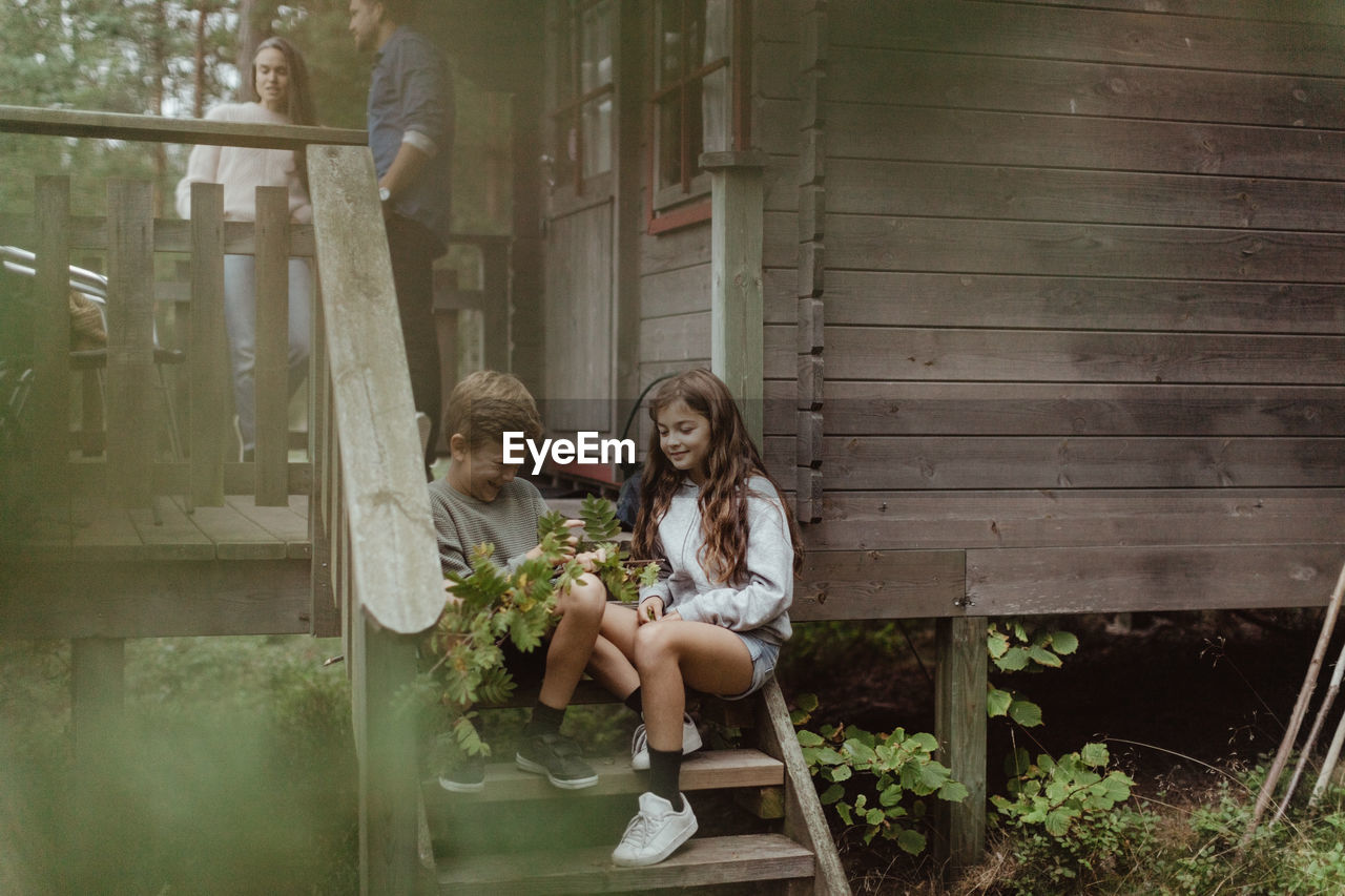 Full length of sibling sitting on doorsteps while parents standing in background