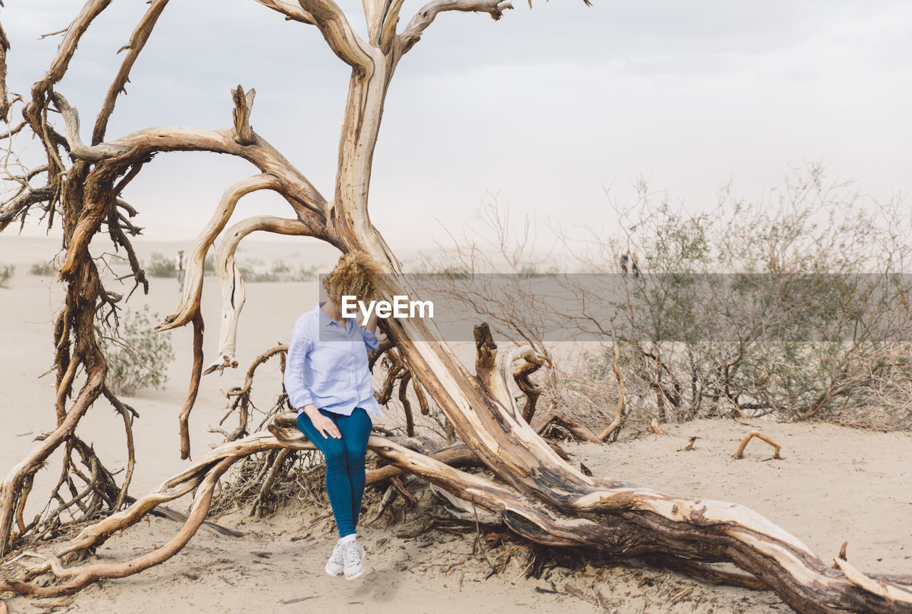 Young woman sitting on fallen tree against cloudy sky at death valley national park