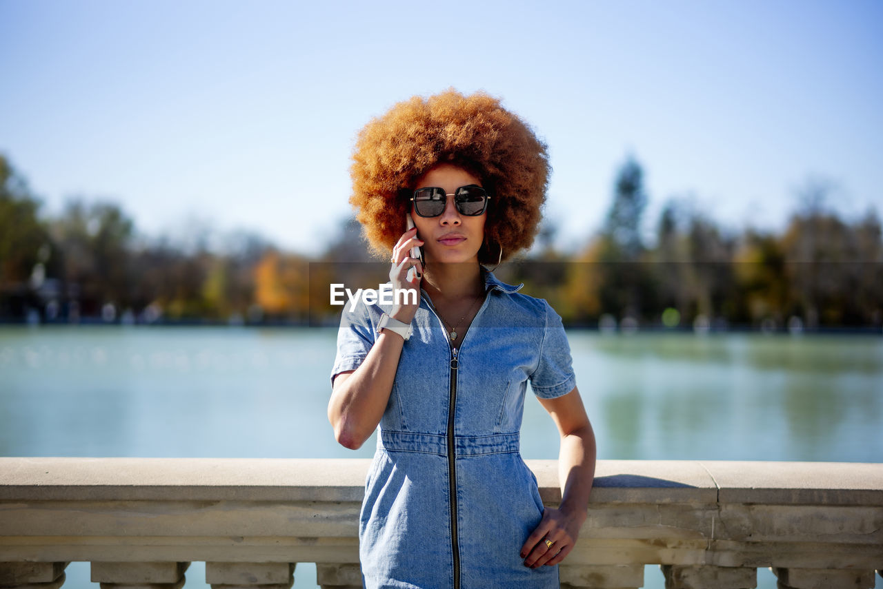 Afro hair woman wearing sunglasses talking on phone at estanque grande del retiro pond on sunny day, spain