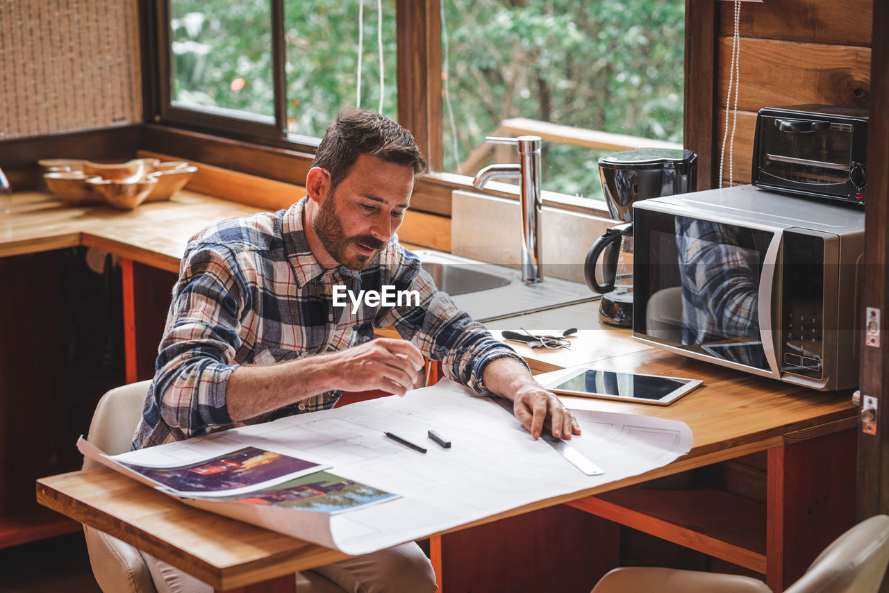 Serious male architect sitting at table in kitchen and drawing blueprint of building with pencil and ruler while working at home