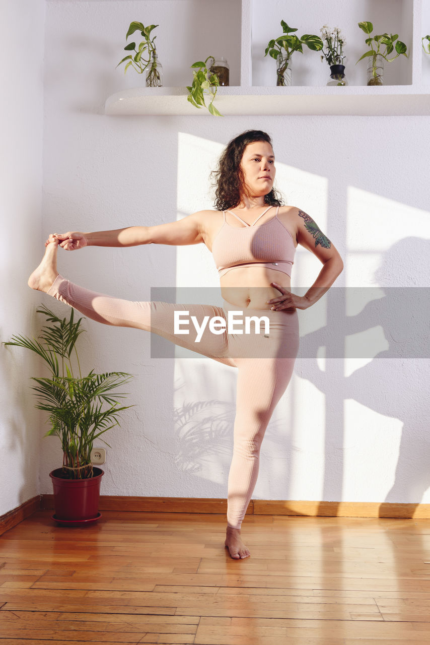 Female in sportswear standing in hasta padangusthasana with raised leg and looking at camera while practicing yoga at home