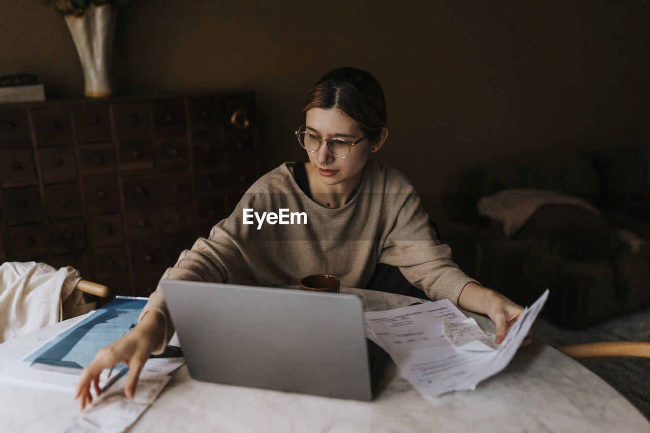 Non-binary person collecting bills while sitting with laptop at home