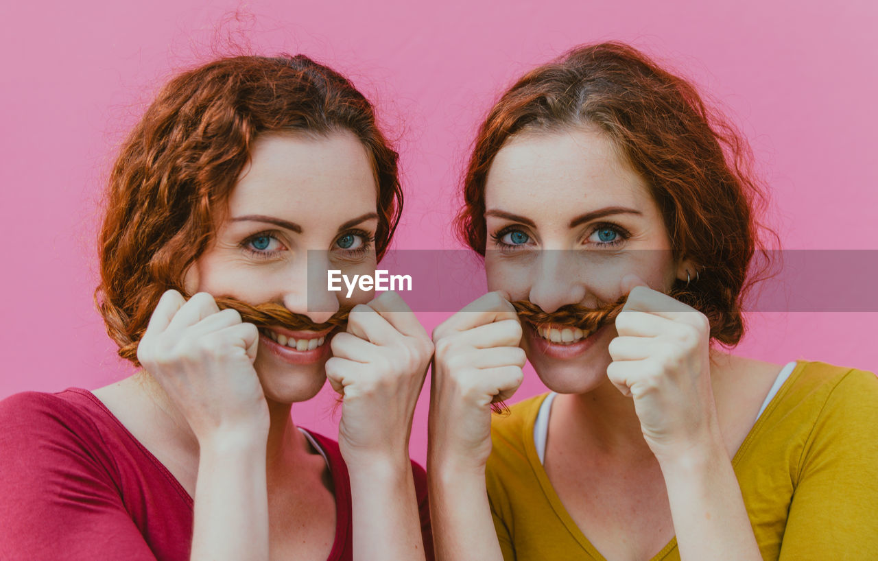 Portrait of smiling siblings making mustache with hair against pink background