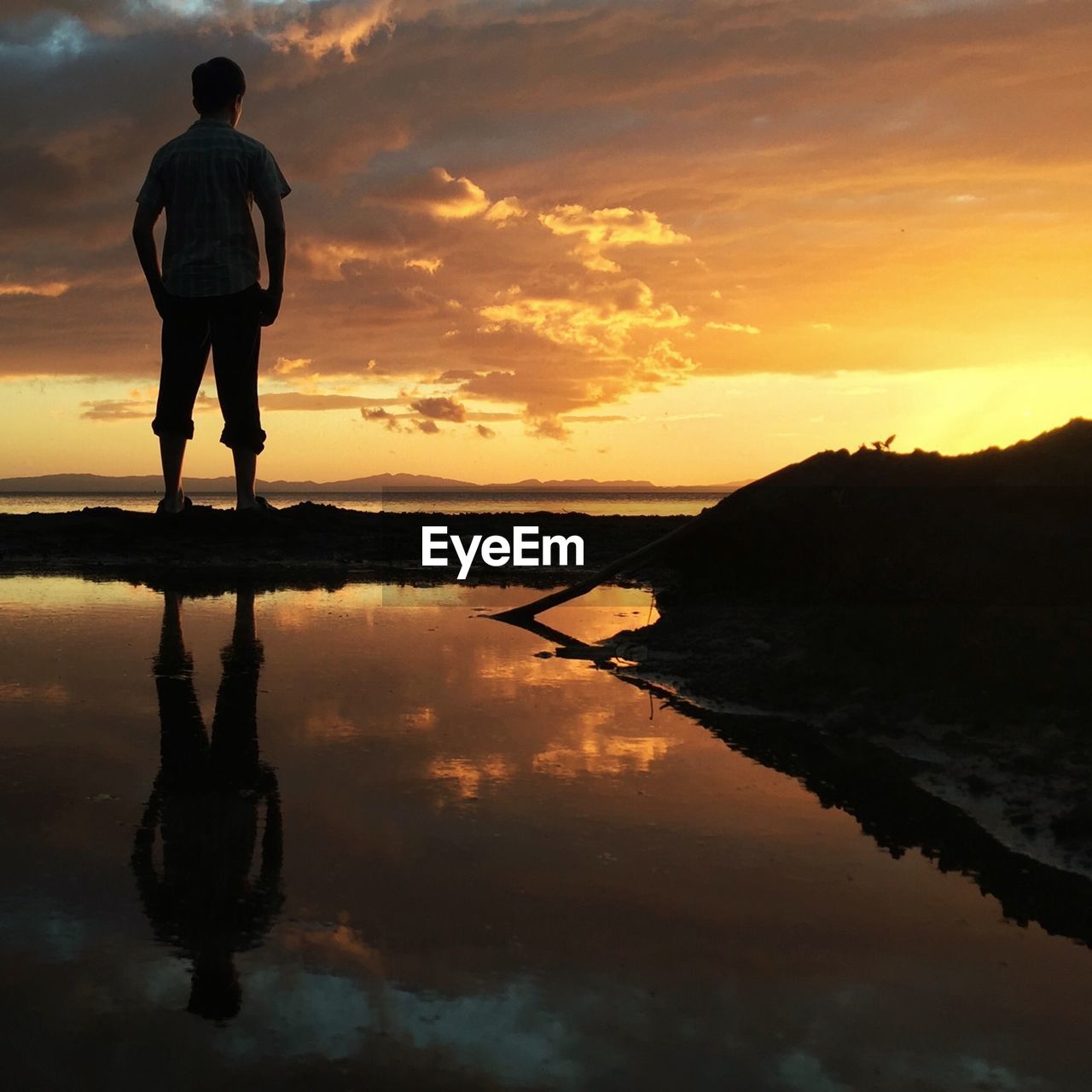 Silhouette man standing at lakeshore against cloudy sky during sunset