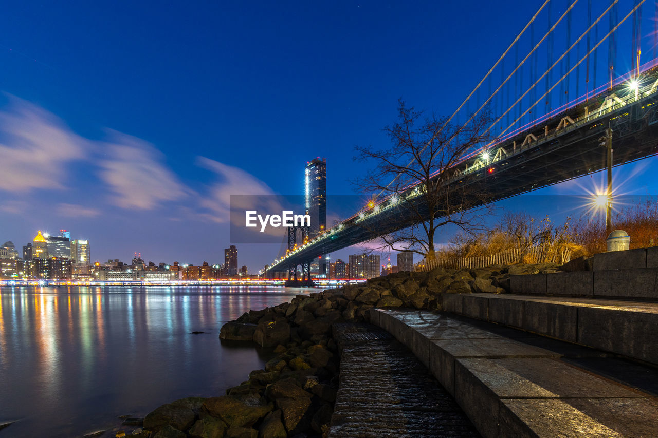 Illuminated bridge over river by buildings against sky at night