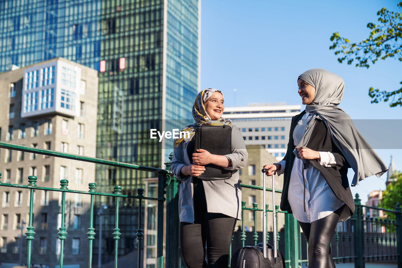 Smiling muslim female entrepreneurs with suitcase walking along street in city and looking at each other