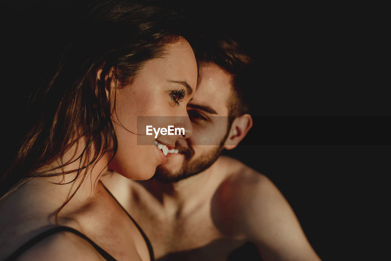 Smiling young couple in swimwear and with wet hair cuddling while resting on dark beach at sunset together during romantic holidays