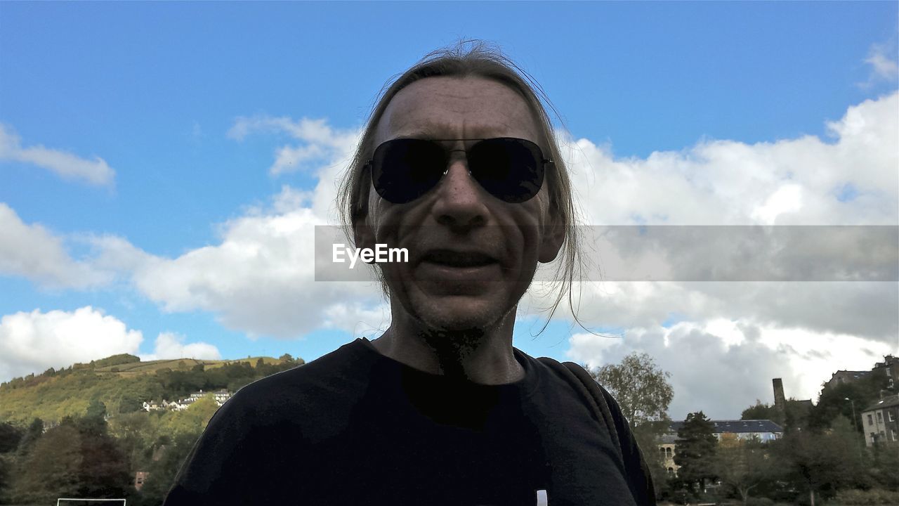 Close-up of mature man wearing sunglasses standing against cloudy sky