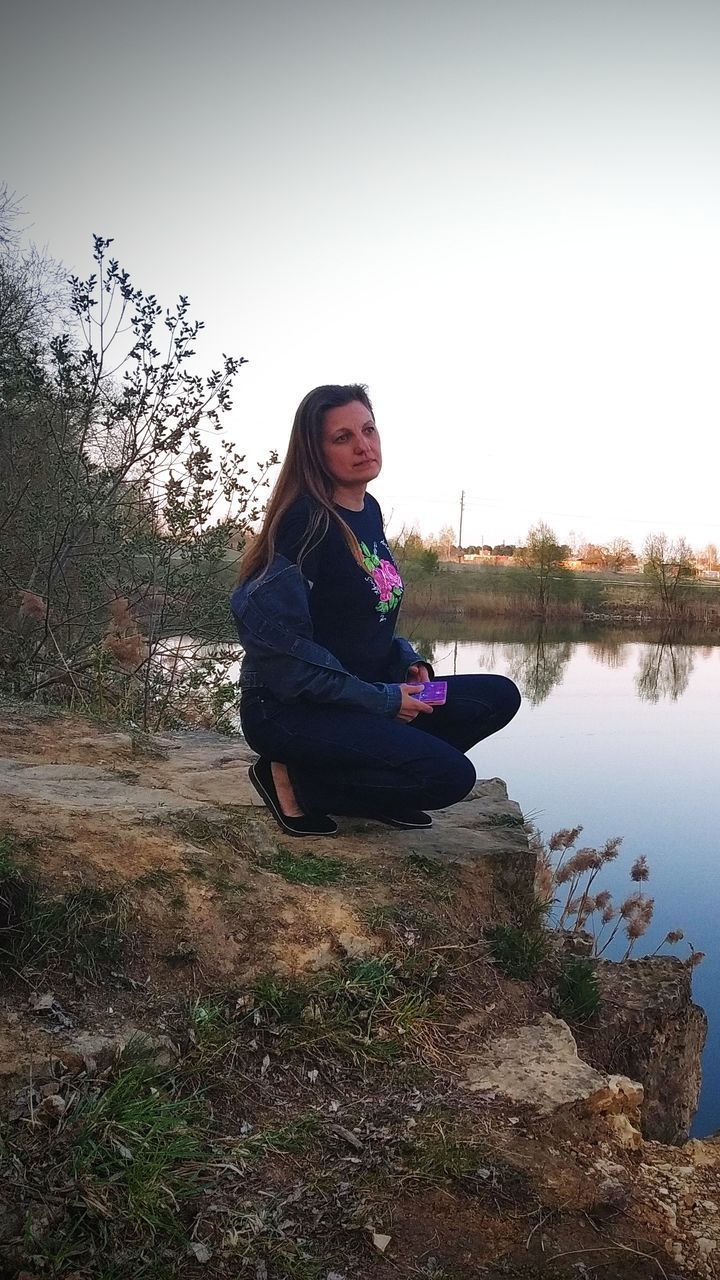 YOUNG WOMAN SITTING BY LAKE AGAINST SKY
