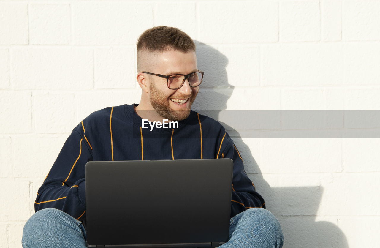 Smiling man using laptop while sitting by wall