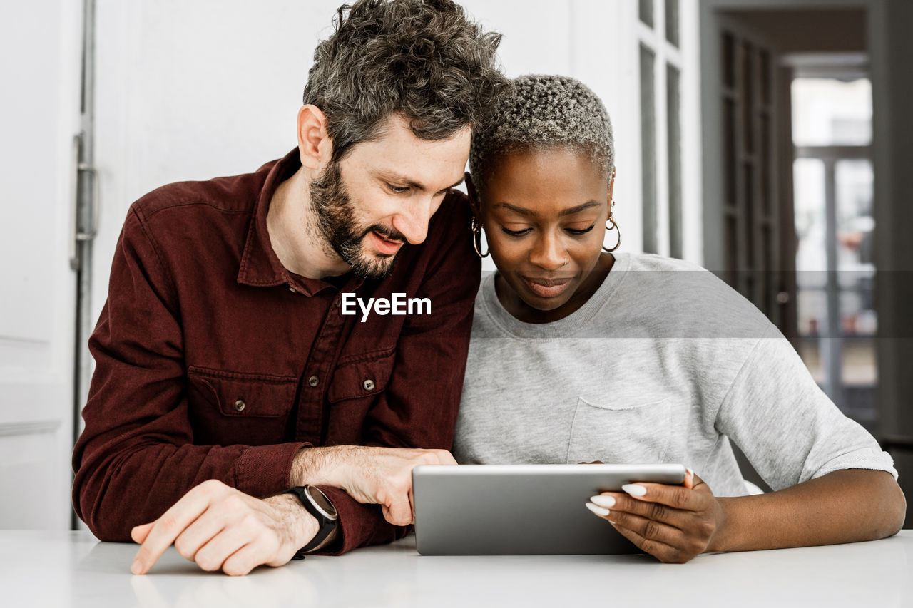 Satisfied african american woman and bearded man smiling while sitting at table and watching digital tablet together at home