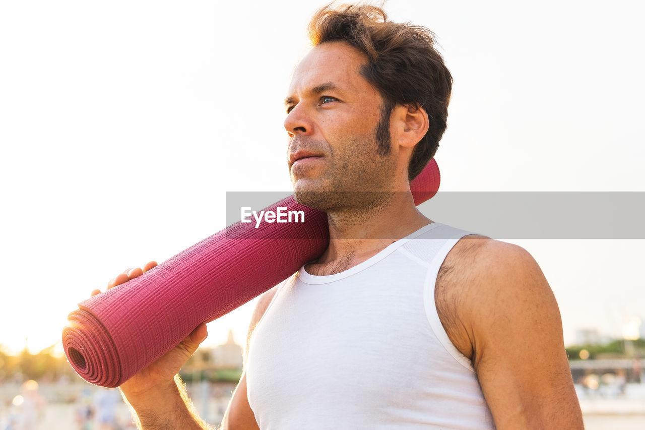 Calm middle aged male carrying rolled up mat on shoulder and looking away during yoga session at sunrise on beach