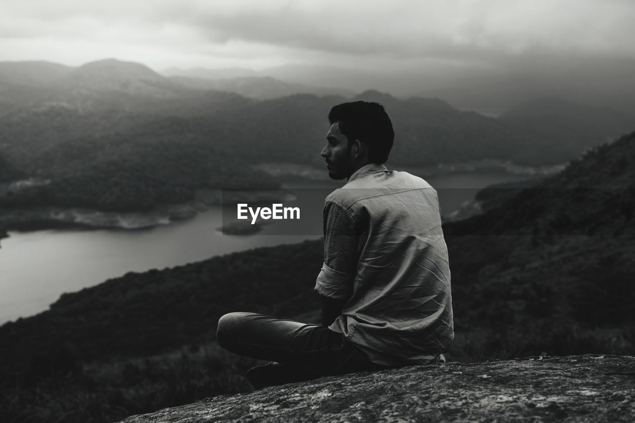 MAN SITTING ON ROCK AGAINST MOUNTAINS