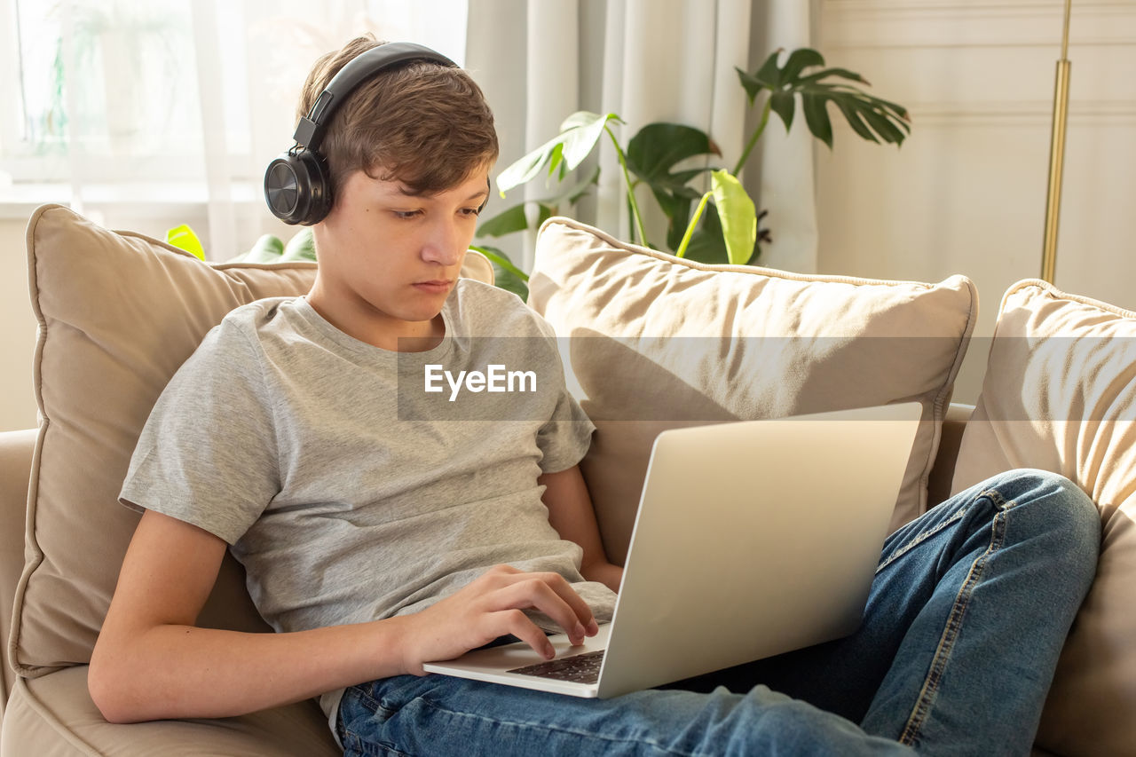 Portrait of teenage , sits on a sofa , wearing black headphones on his head, using a laptop.