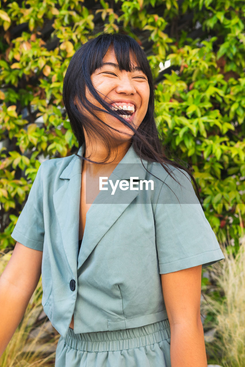 Cheerful young ethnic female in stylish clothes laughing with closed eyes against lush bush on sunny summer day in park