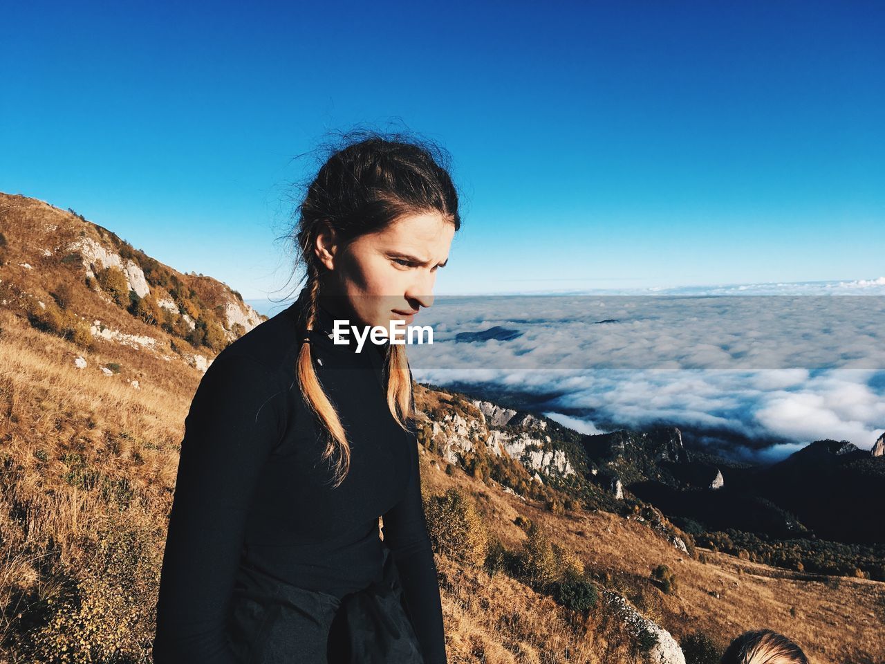 Young woman standing on cliff by cloudscape against clear sky