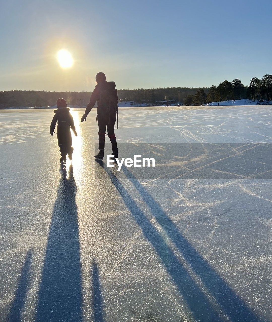 winter, snow, sunlight, cold temperature, sky, nature, shadow, two people, full length, men, adult, ice, sports, togetherness, winter sports, ice skating, child, lens flare, back lit, leisure activity, family, childhood, footwear, day, sunny, lifestyles, father, landscape, freezing, parent, women, female, frozen, environment, clear sky, blue, cloud, one parent, bonding, copy space, mature adult, sun, land, reflection, positive emotion, warm clothing, skiing, sunset, outdoors, walking