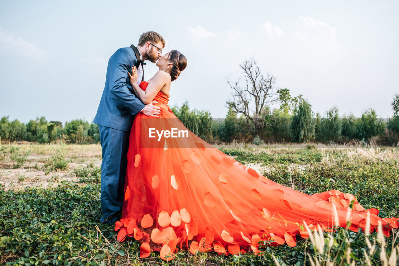 Newlywed couple kissing on field against sky